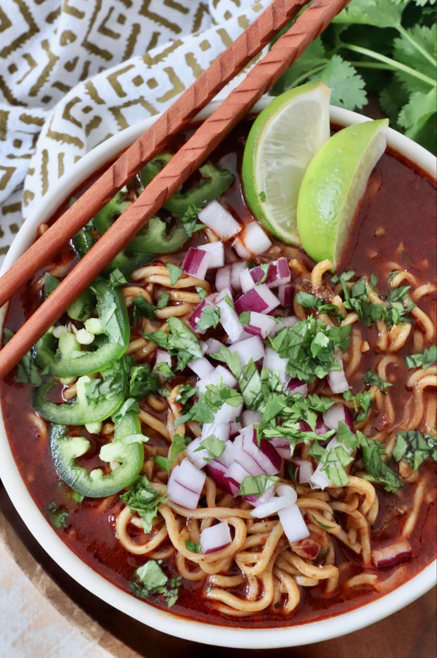 A finished birria ramen bowl with a lime wedge and a small cup of consommé on the side